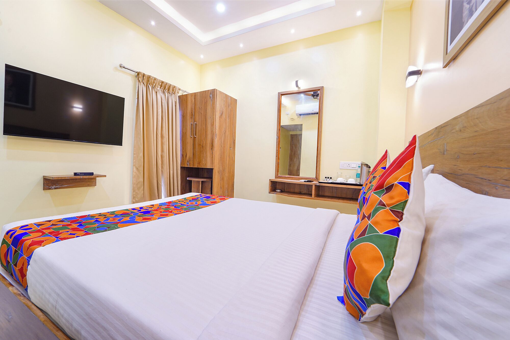 A hotel room with a large bed, a TV mounted on the wall, a wooden wardrobe, a mirror, and a bedside table with a lamp.