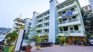 A multi-story hotel with a sign reading "Shalom Indus Residency" and a "For Hotels" sign. There are outdoor seating areas with tables and chairs, and potted plants. - Fabhotel Shalom Indus Residency (Candolim)
