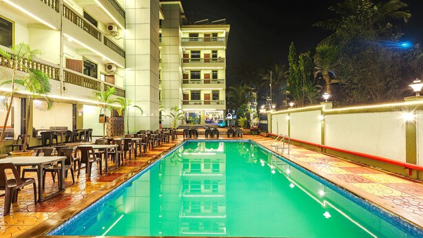 A swimming pool with a tiled deck, surrounded by tables and chairs, and a multi-story building in the background. - Fabhotel Shalom Indus Residency (Candolim)