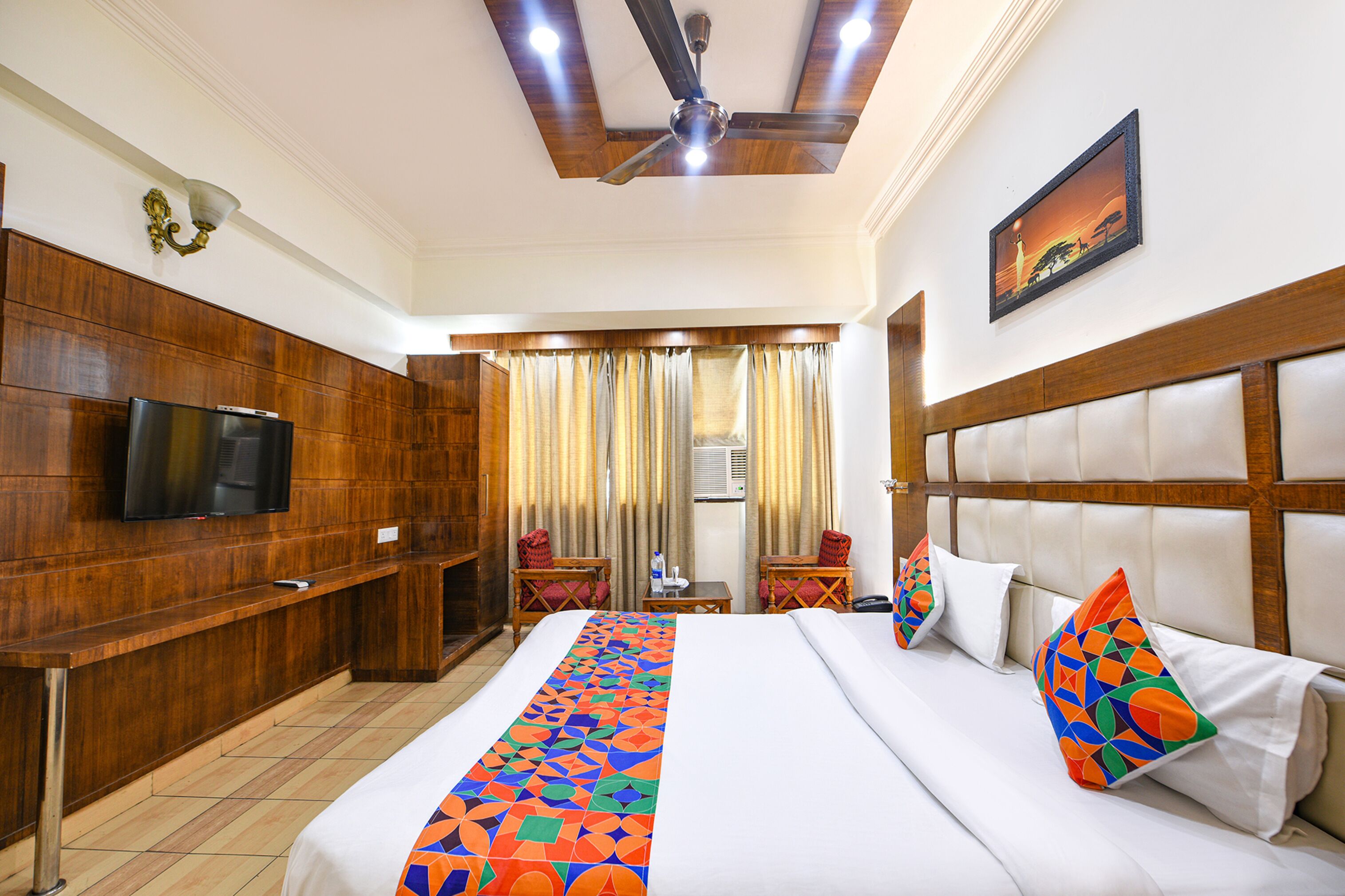 A hotel room with a large bed, a television, wooden paneling, a ceiling fan, and a painting on the wall.