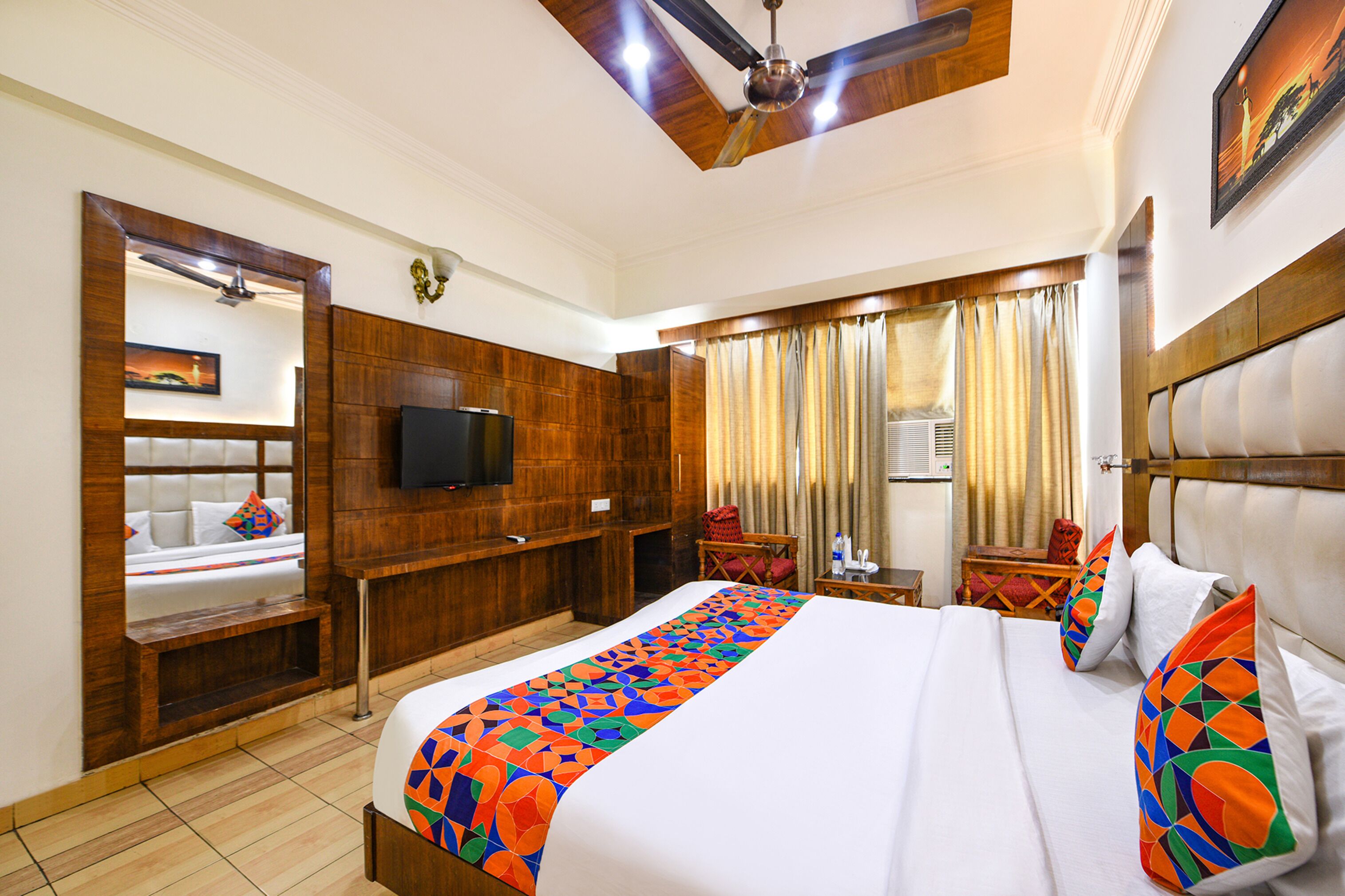 A hotel room with a large bed, wooden headboard, a flat-screen TV, a ceiling fan, and a window with curtains.