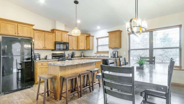 A modern kitchen with wooden cabinets, a stainless steel refrigerator, a central island with stools, and a dining area with a chandelier.