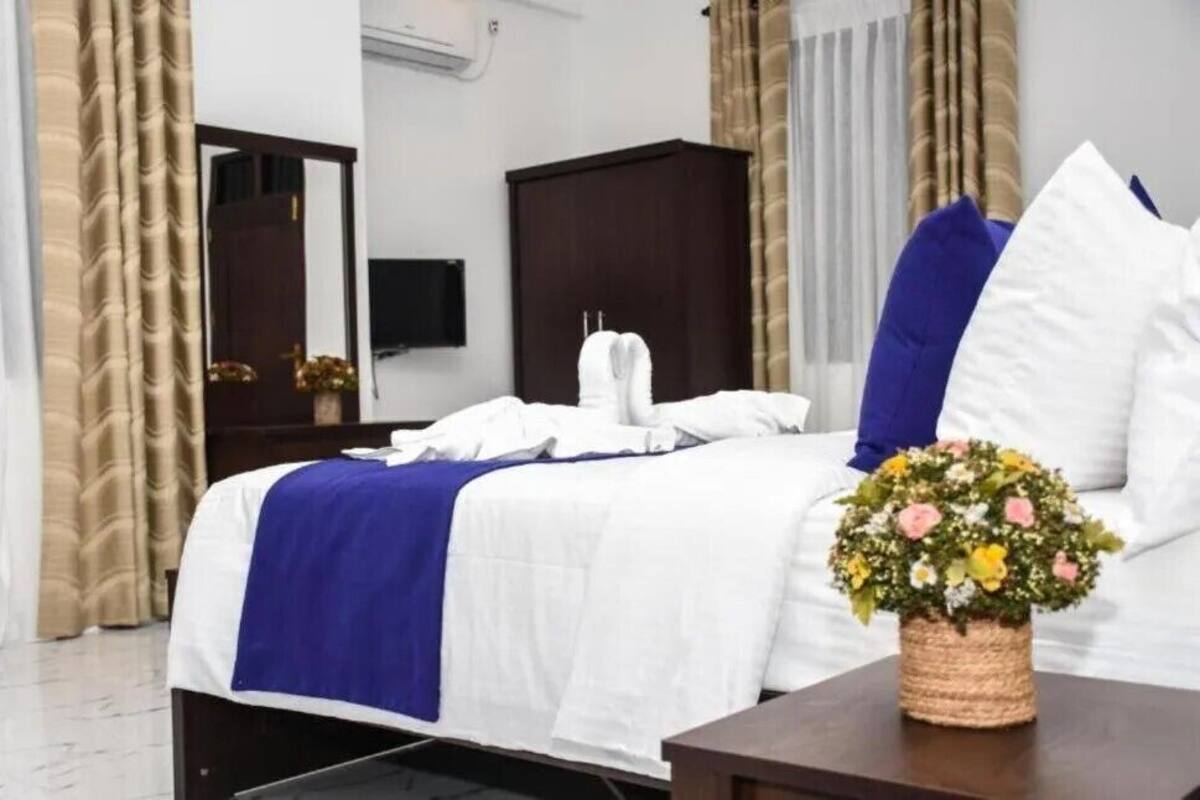A hotel room with a neatly made bed, a dark wooden wardrobe, a flat-screen TV mounted on the wall, and a decorative flower arrangement on the nightstand.