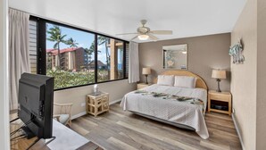 Ein Hotelzimmer mit Bett, Schreibtisch mit Computer, Deckenventilator und Blick auf Palmen und Gebäude.