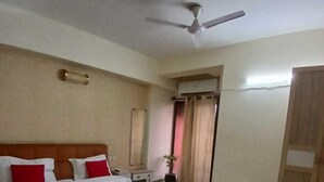 A bedroom with a bed, a red rug, a ceiling fan, a wardrobe, and a small table with a plant. - Shree Krishna Valley (Vrindavan)