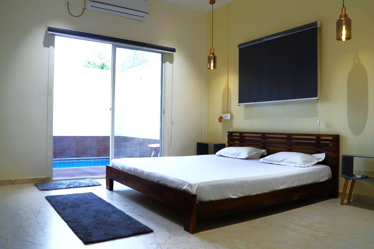 A bedroom with a large bed, a sliding glass door to a balcony, a mounted TV, and a hanging pendant light.