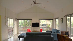 A spacious living room with a large grey sofa, a television mounted on the wall, and a ceiling fan. - Mango Shack Villa (Trimbak)