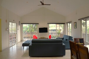 A spacious living room with a large grey sofa, a television mounted on the wall, and a ceiling fan.
