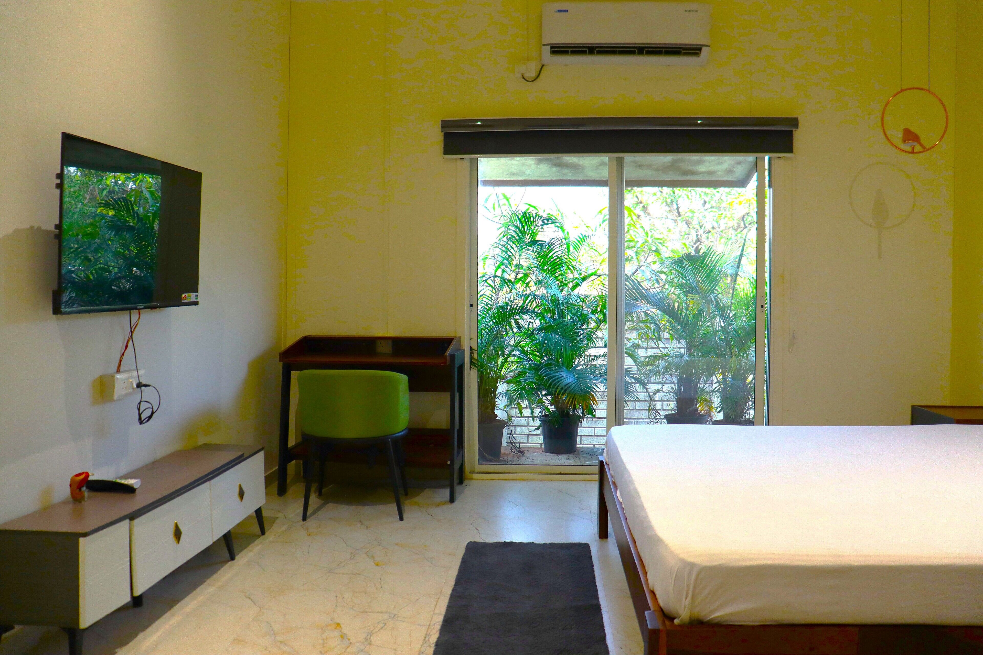A bedroom with a bed, a desk, a chair, a television, and a window with a view of greenery.