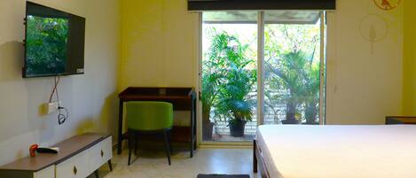 A bedroom with a bed, a desk, a chair, a television, and a window with a view of greenery.