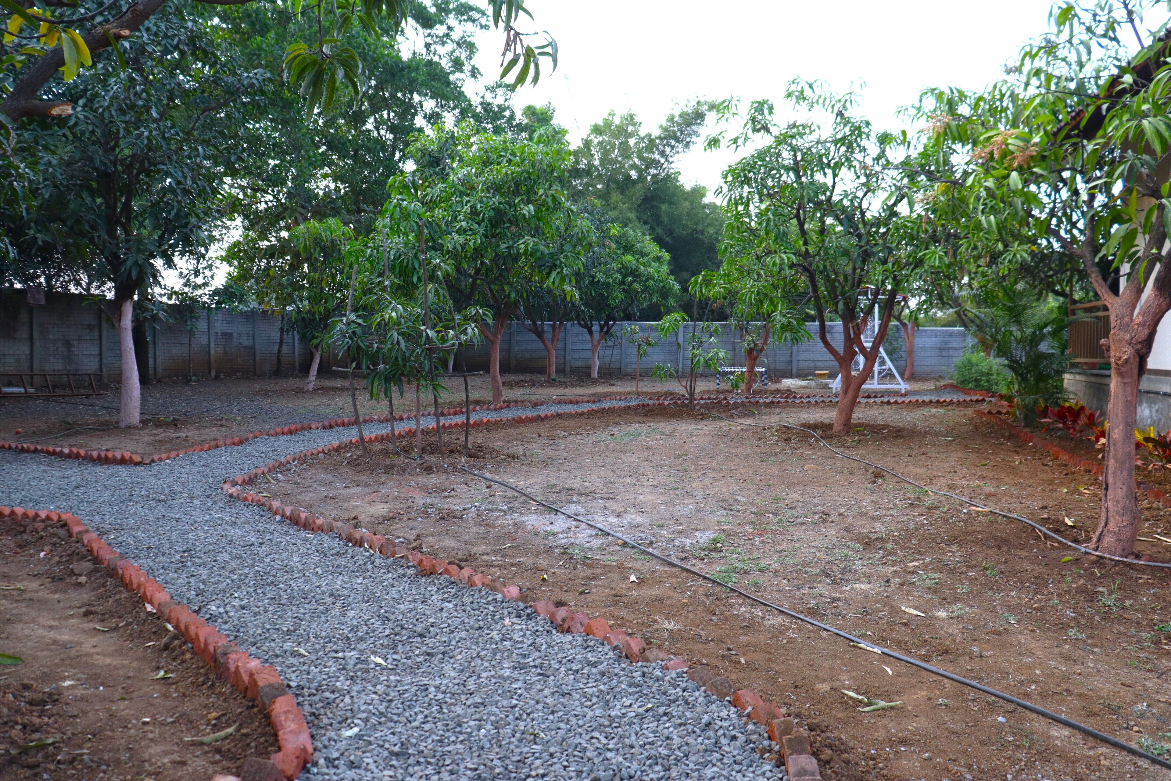 Un jardín con senderos de grava y árboles, un muro de ladrillo y una valla metálica.