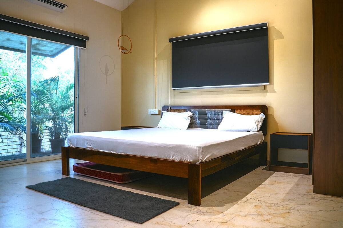 A bedroom with a bed, a nightstand, a window with a view of palm trees, and a wall-mounted TV.