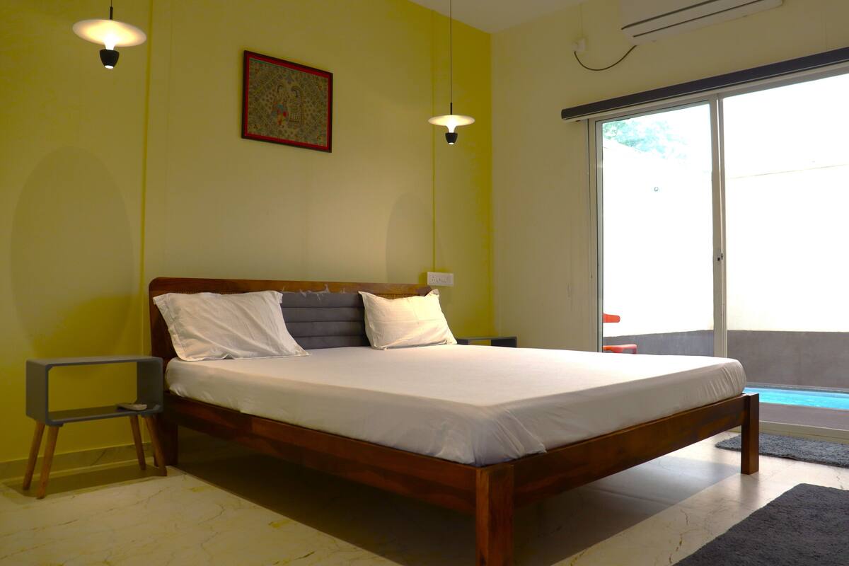 A bedroom with a wooden bed, white bedding, a small bedside table, and a large window with a view of a swimming pool.