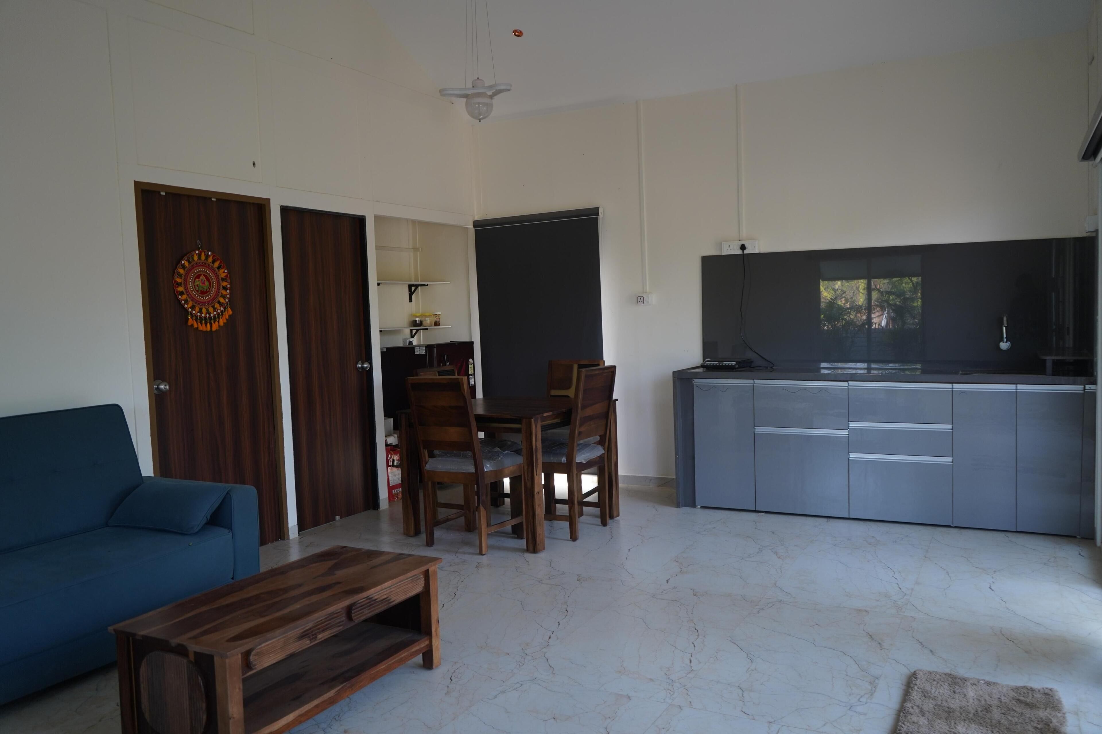 A modern living room with a blue sofa, wooden coffee table, and a dining area with a table and chairs. There is a kitchen area with grey cabinets and a TV.