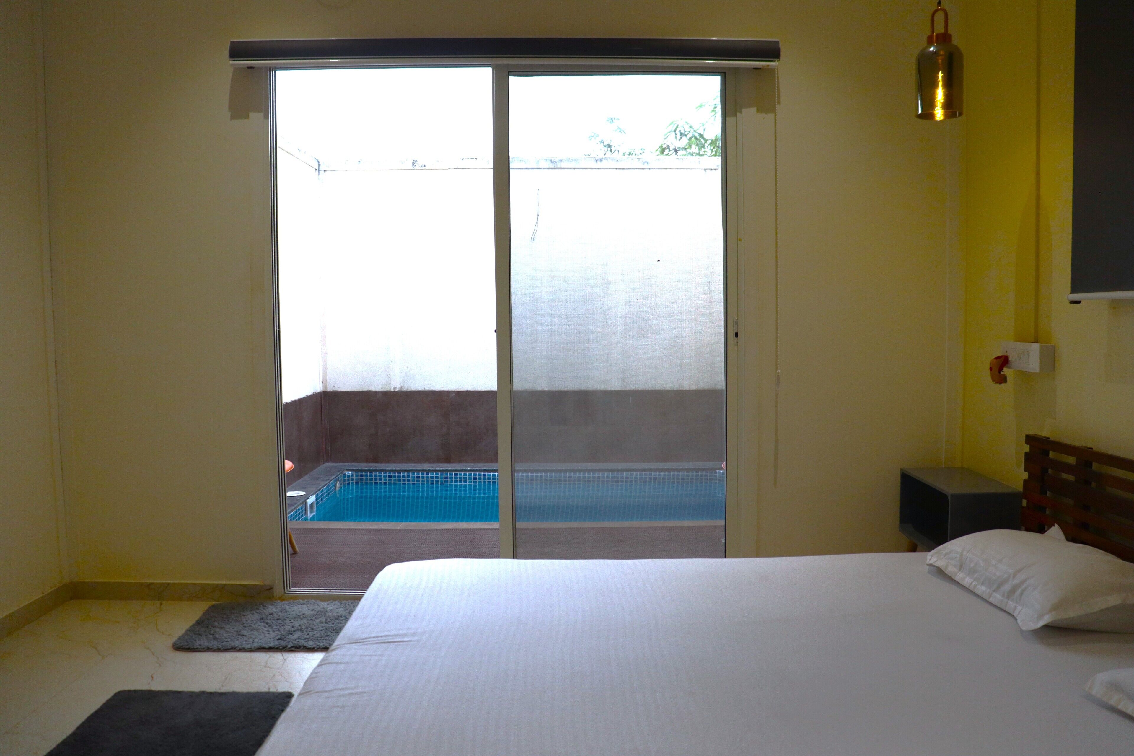 Un dormitorio con una cama grande, un ventanal corredero que da a un balcón con piscina y una mesita de noche con lámpara.