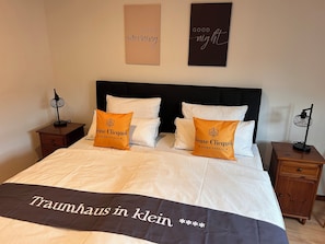 Una cama bien hecha con ropa blanca y dos almohadas naranjas, un cabecero negro y dos mesitas de noche con lámparas.