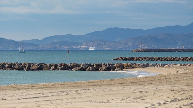 A beach with rocky breakwaters, calm sea, and distant mountains.
