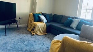 En stue med et stort fladskærms-tv, en mørk sofa med mange puder og et gult pled.