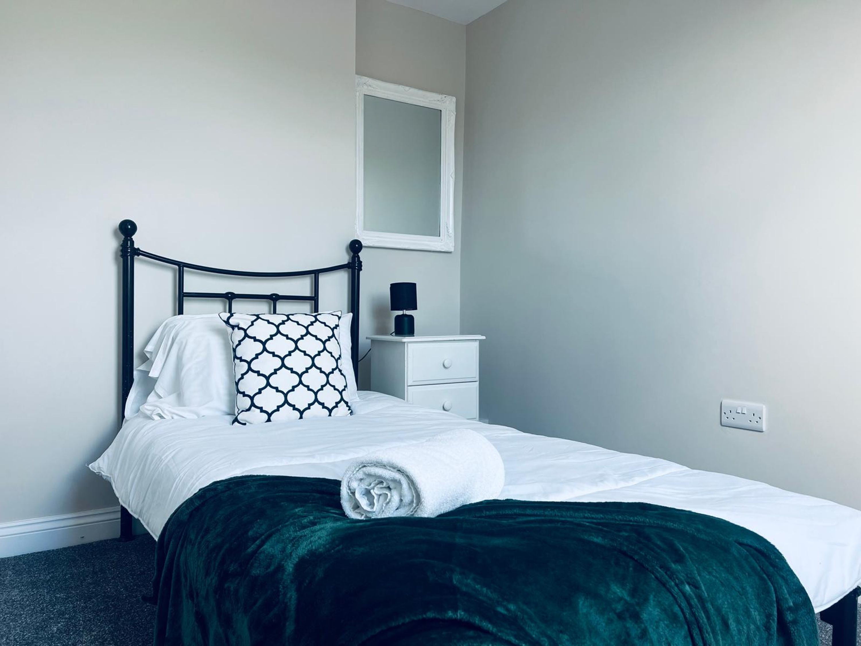 A neatly made bed with a dark green blanket, a white pillow with a black pattern, and a white pillow. There is a small bedside table with a lamp and a mirror above the bed.