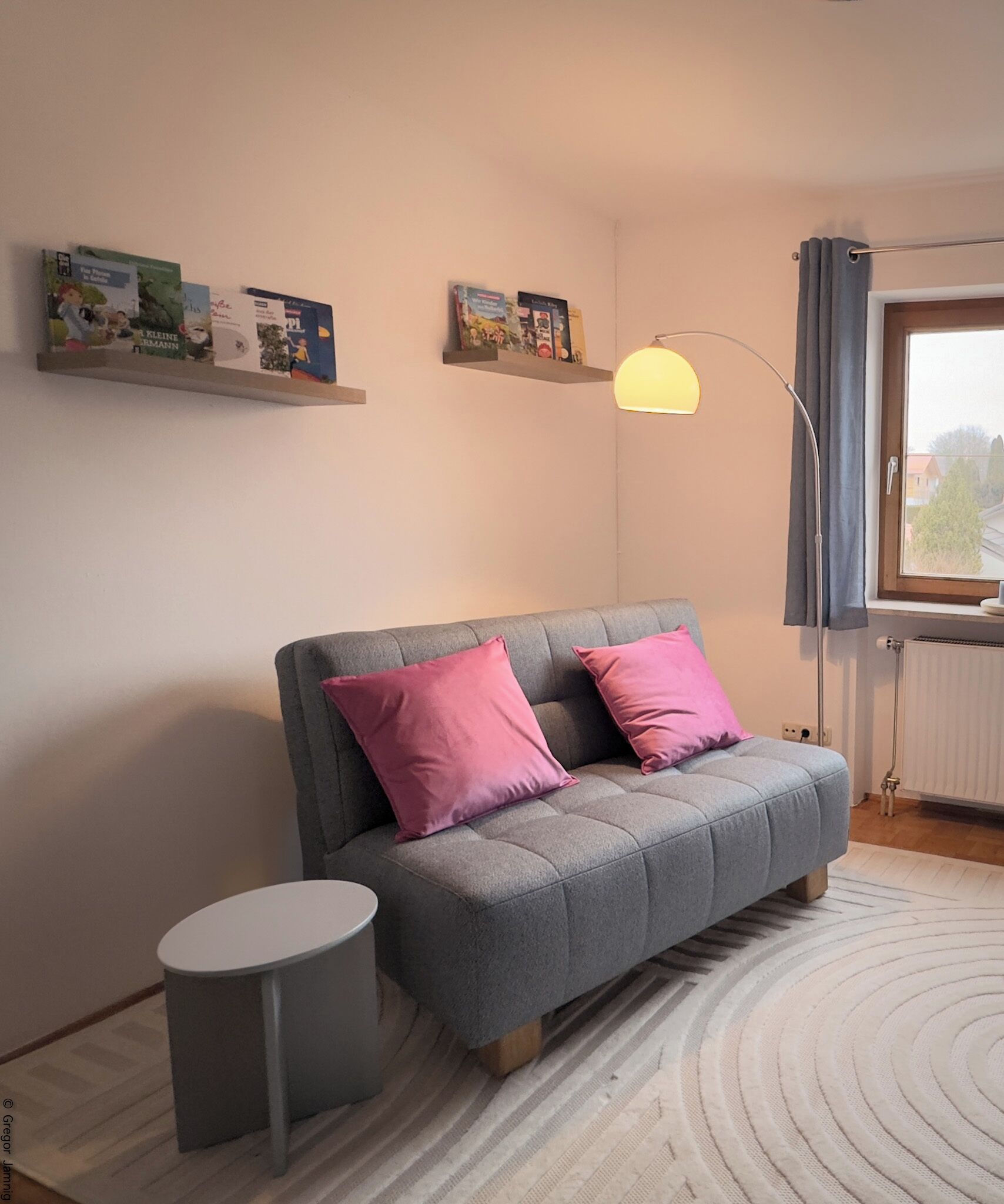 A modern living room with a grey sofa, two pink cushions, a small round side table, and a floor lamp. There are bookshelves above the sofa with books and decorative items.