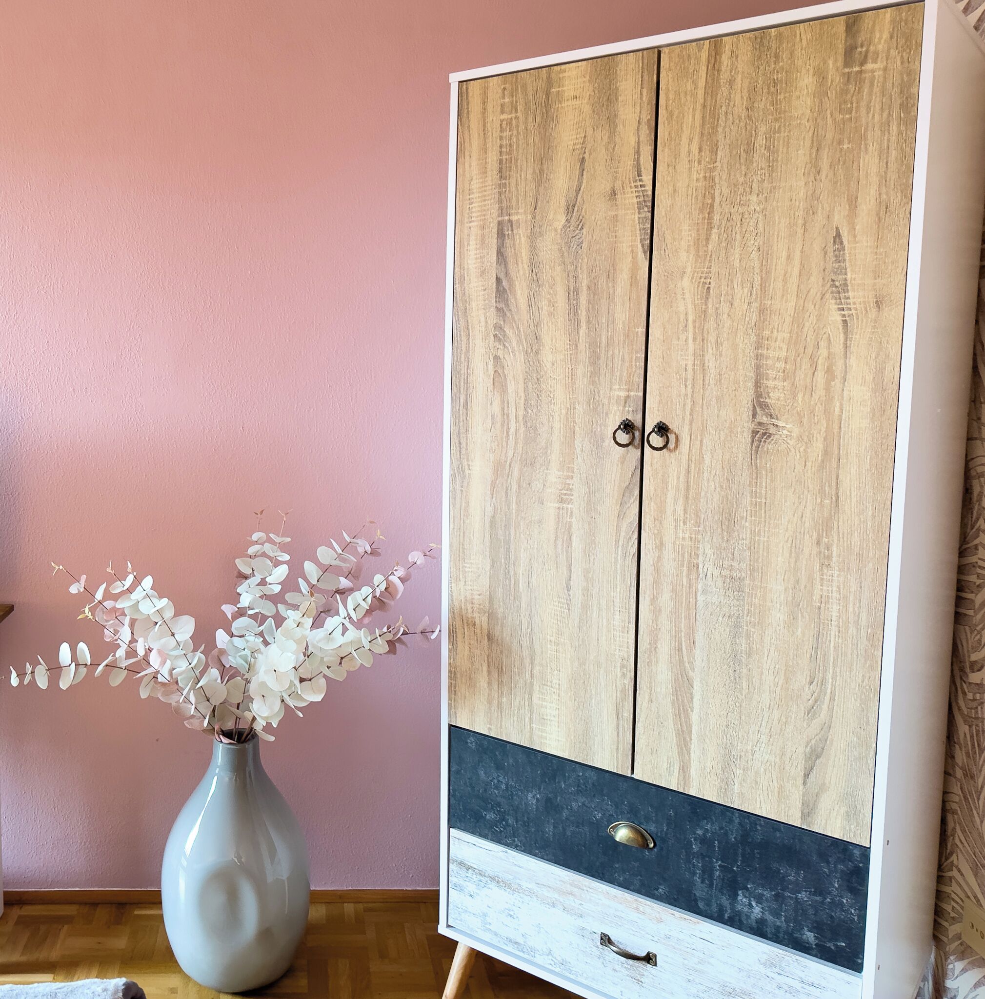 A wooden wardrobe with two doors and a lower drawer, a vase with white flowers, and a pink wall.