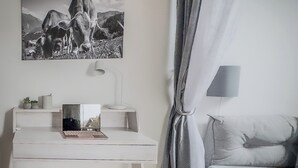Ein schlicht eingerichteter Raum mit einem weißen Schreibtisch, einem Laptop, einer Lampe, einem Bett und einem großen Gemälde von Bergen.
