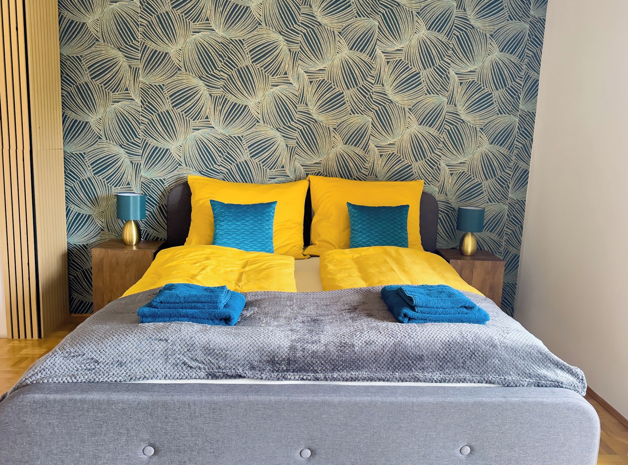 Een netjes opgemaakt bed met gele en blauwe beddengoed, twee bedlampen en behang met een patroon.