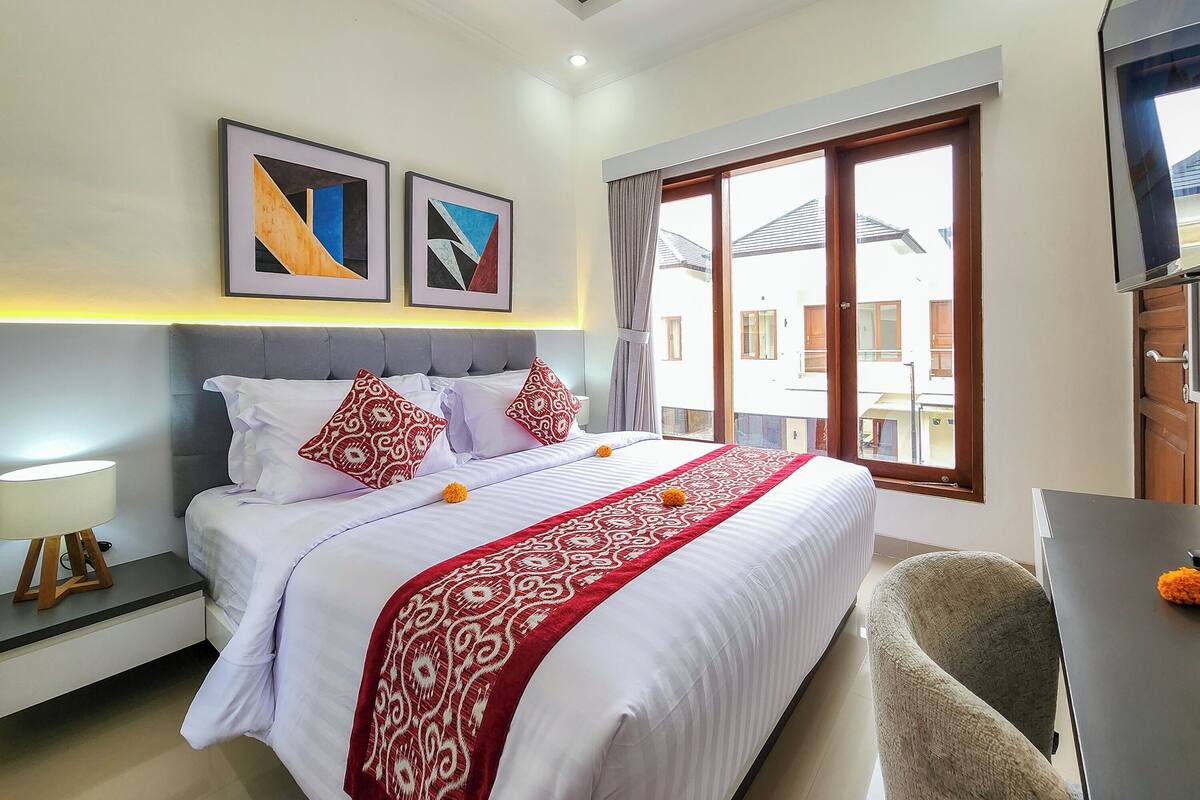 A hotel room with a large bed, two pillows, a red and white patterned bedspread, a nightstand with a lamp, a chair, and a TV on a stand.
