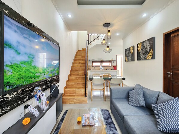 A modern living room with a large flat-screen TV, a grey sofa, a coffee table, and a staircase leading to the kitchen. - Villa Elemi Seminyak  by Nagisa Bali (Seminyak)