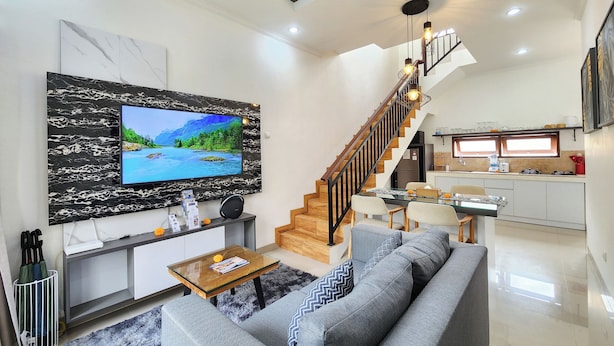 A modern living room with a large flat-screen TV, a grey sofa, a wooden staircase, and a kitchen area.