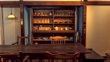 Un comedor con una mesa de madera larga y sillas, una estantería con distintos vasos y platos, y una lámpara colgante.