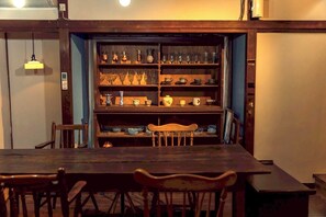 Un comedor con una mesa de madera larga y sillas, una estantería con distintos vasos y platos, y una lámpara colgante.