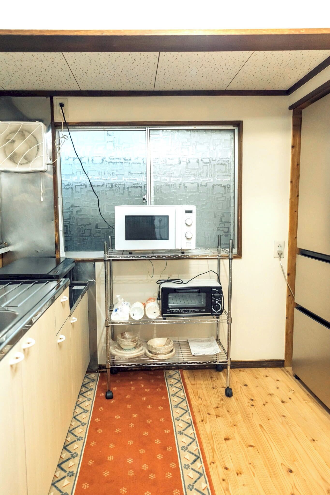 A compact kitchen with a microwave, toaster, and dish rack on a metal stand. A patterned rug is on the floor, and a window with a view of buildings is above the counter.