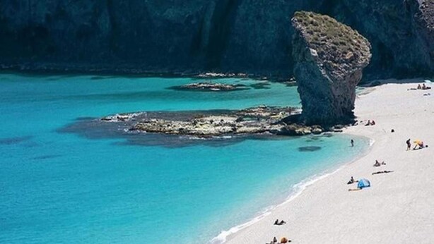 A beach with clear turquoise water, a prominent rock formation, and a sandy shore with people and a tent.