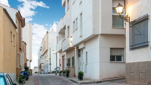 A narrow urban street lined with multi-story buildings, featuring balconies, potted plants, and street lamps.