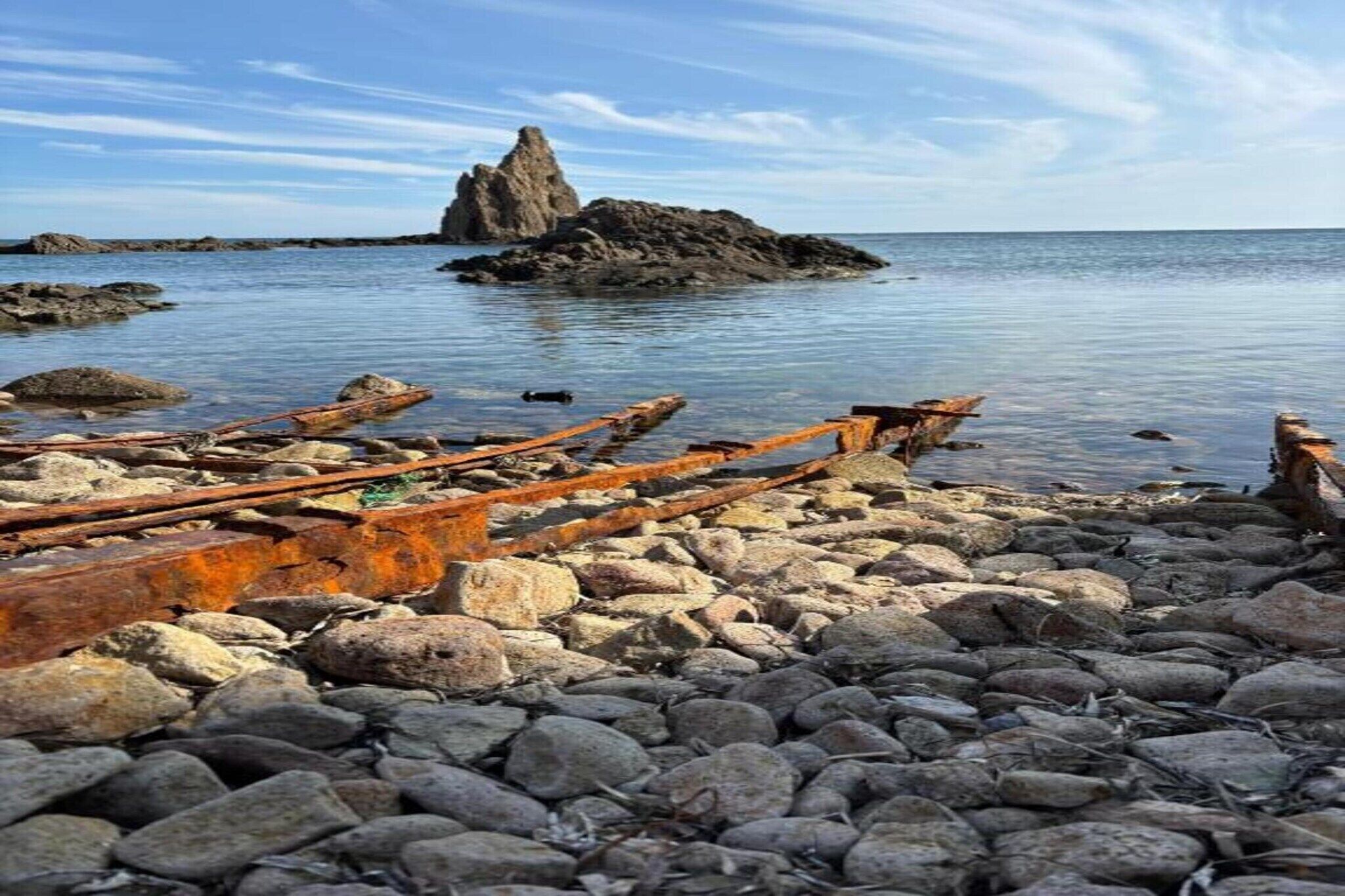 A rocky shoreline with a large rock formation in the distance and rusted metal debris in the foreground.