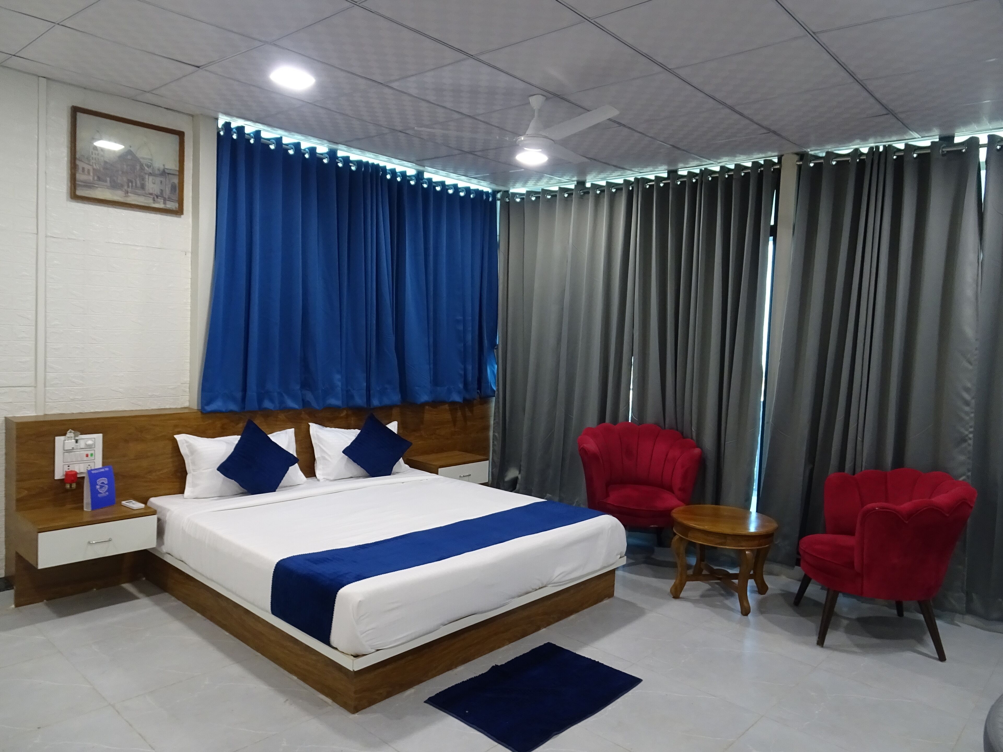 Quarto de hotel com uma cama, uma mesa de cabeceira, uma poltrona vermelha, uma mesinha de madeira e uma pequena mesa redonda.