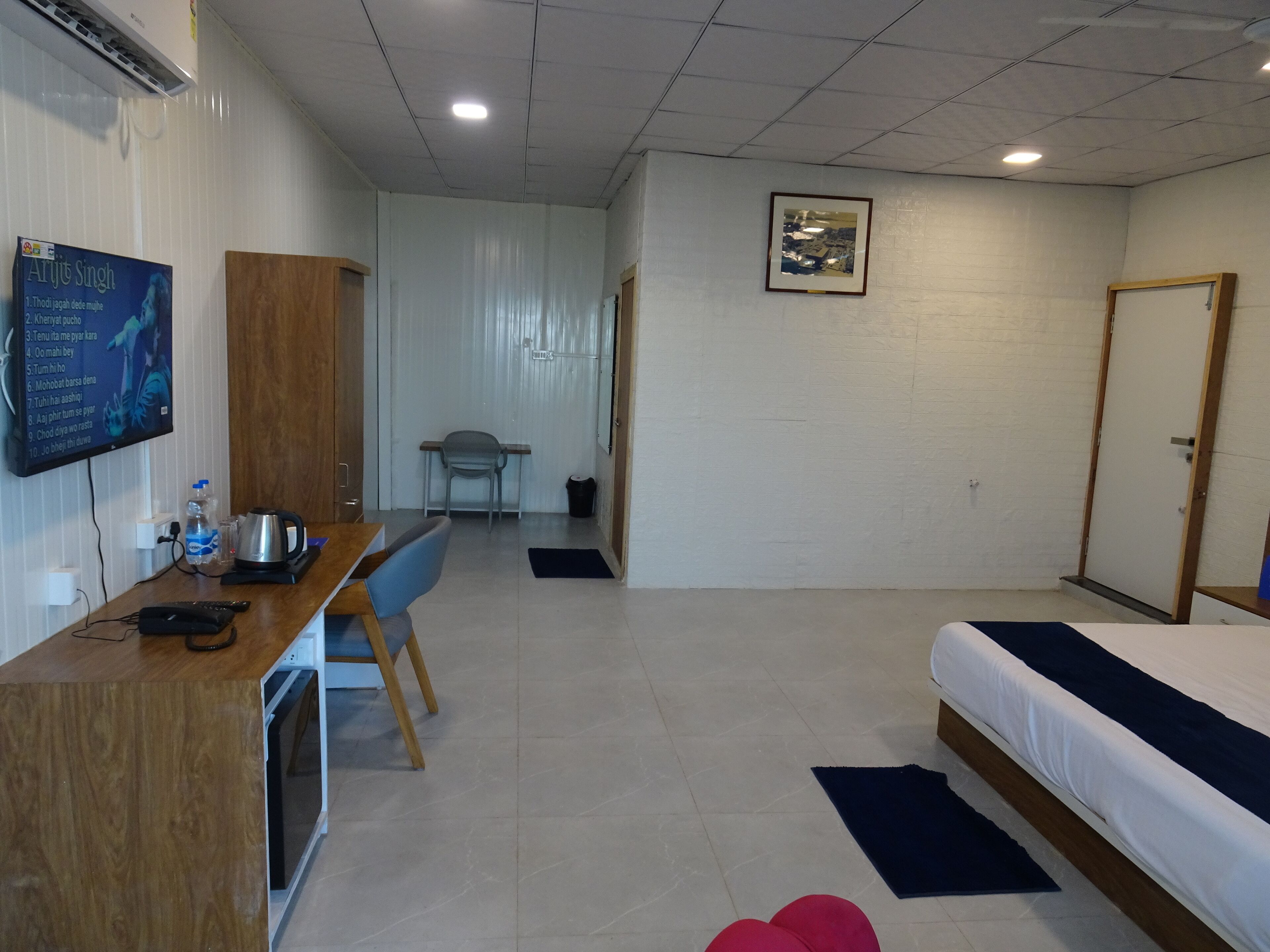 Une chambre d’hôtel avec un bureau en bois, un lit, une chaise, une petite table, un tableau au mur et une porte.