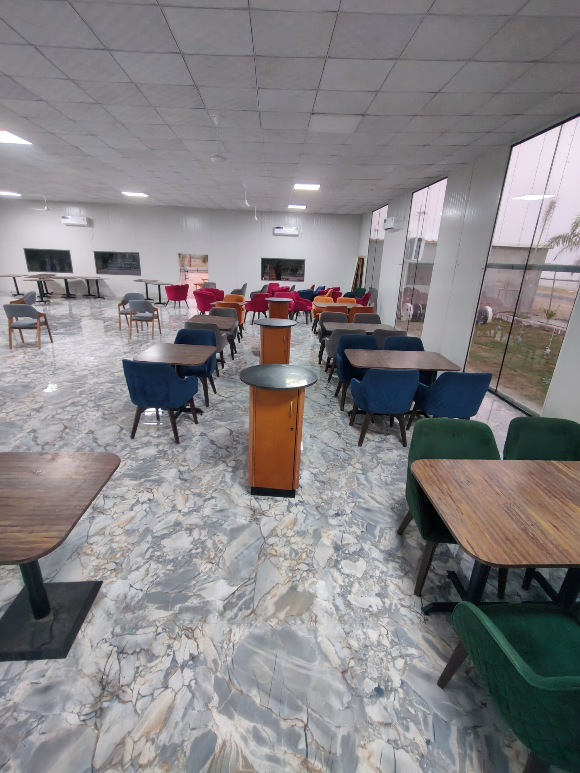 A spacious room with a marble floor, multiple tables and chairs, and large windows offering a view of the outside.