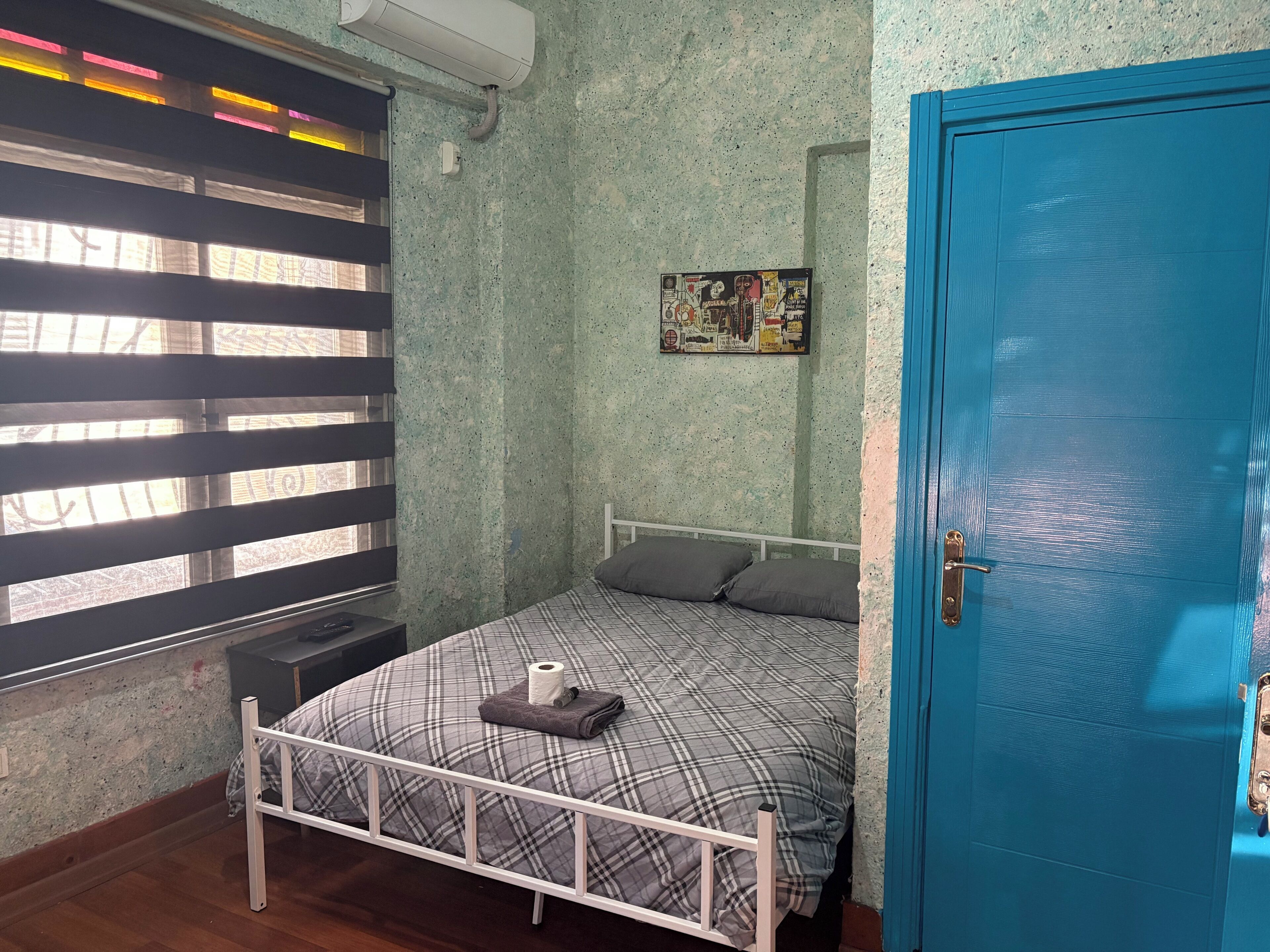 A room with a bed, a blue door, a wall-mounted air conditioner, and a poster.