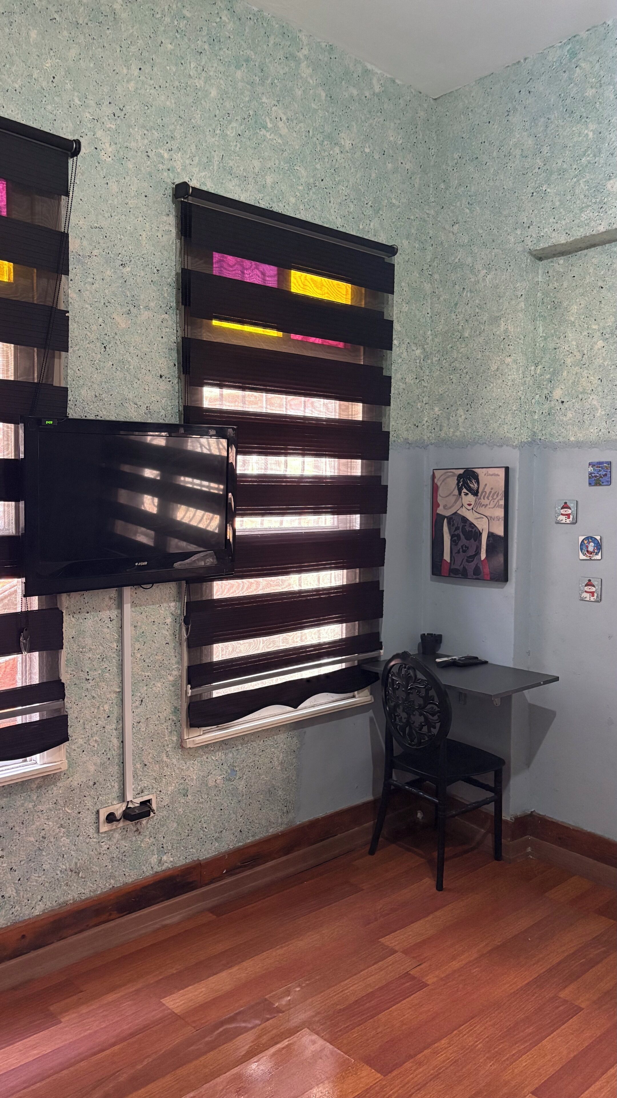 Una habitación con piso de madera, un escritorio con una silla, un televisor montado en la pared y una ventana con persianas.