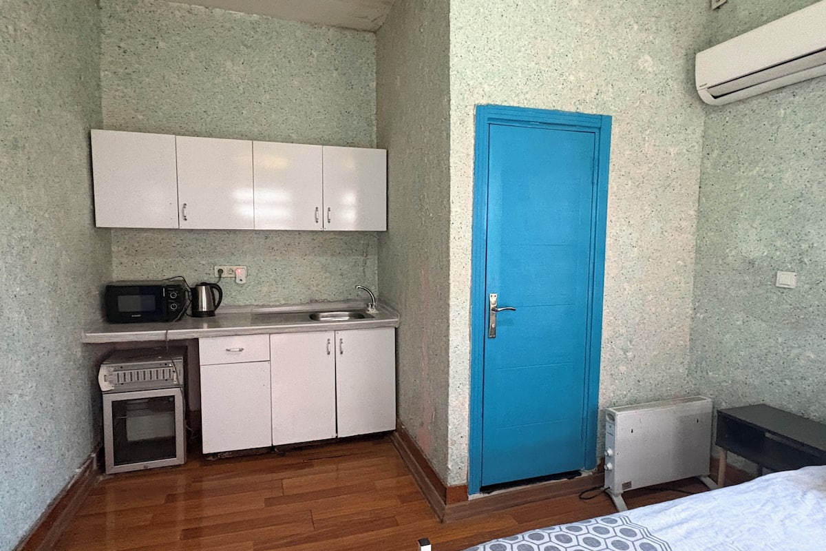 A compact room with a kitchenette, a blue door, and a bed with a patterned bedspread.