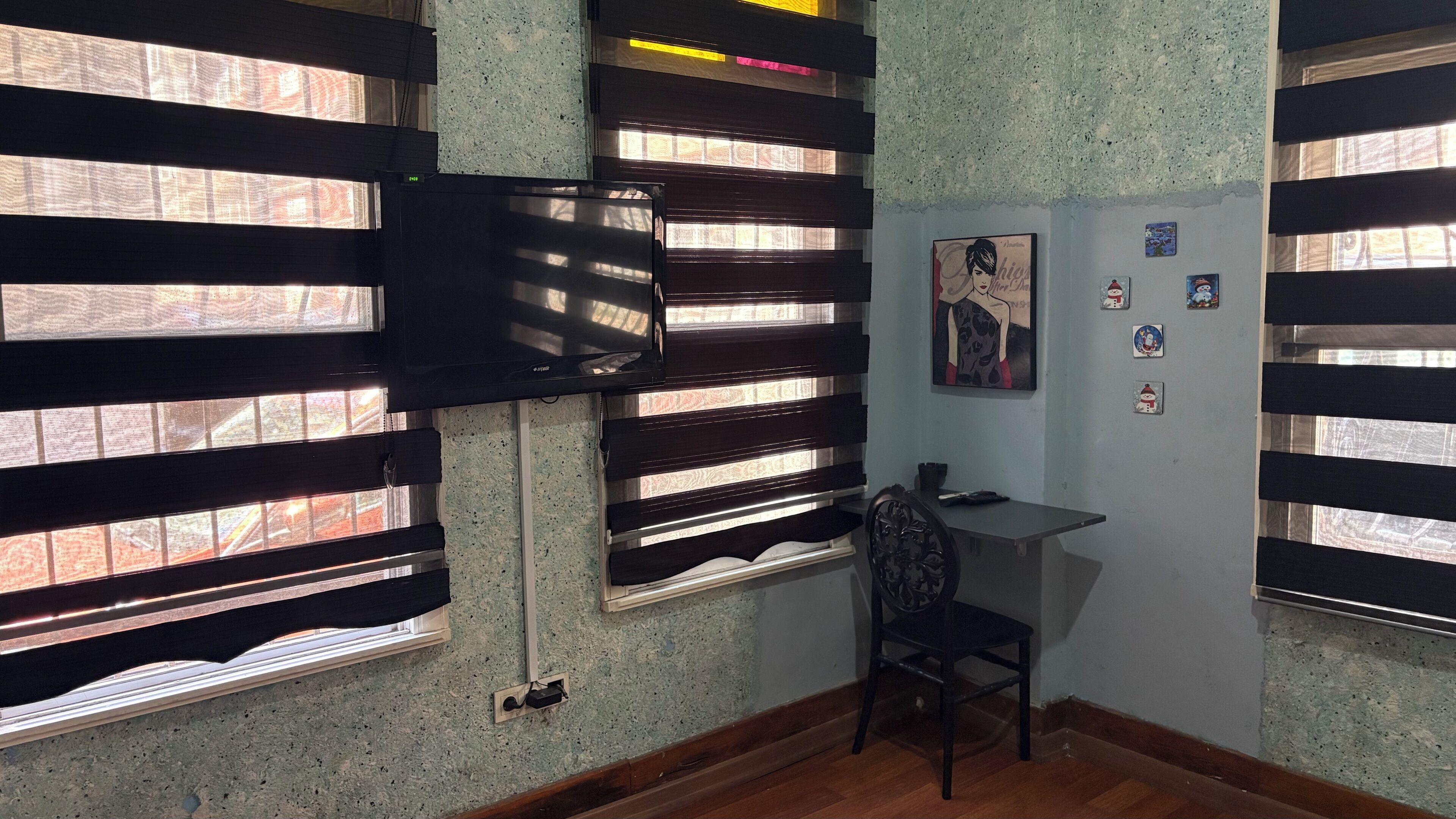A room with a TV mounted on the wall, a desk with a chair, and a window with blinds.
