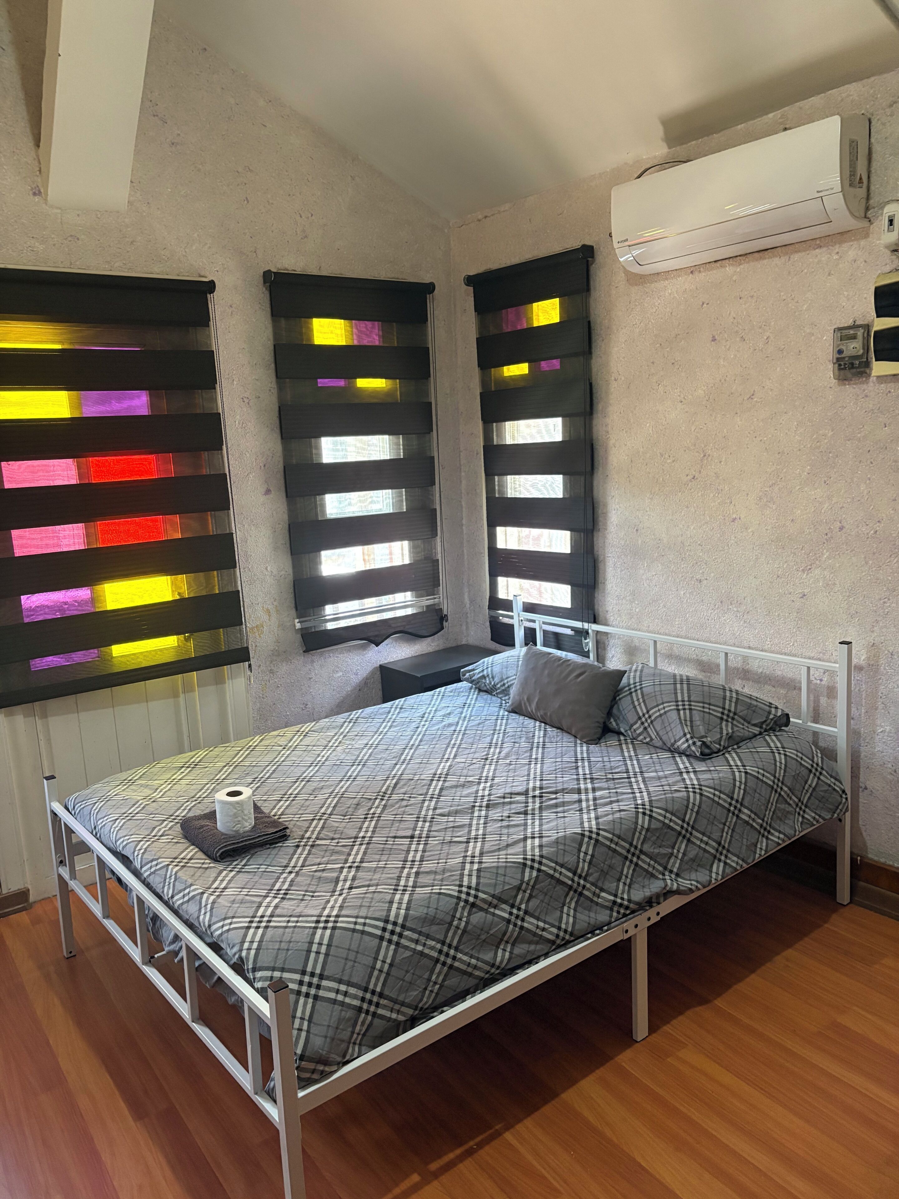A single-bed room with a metal frame, a plaid bedspread, a pillow, a bedside table with a lamp, and a window with colorful blinds.