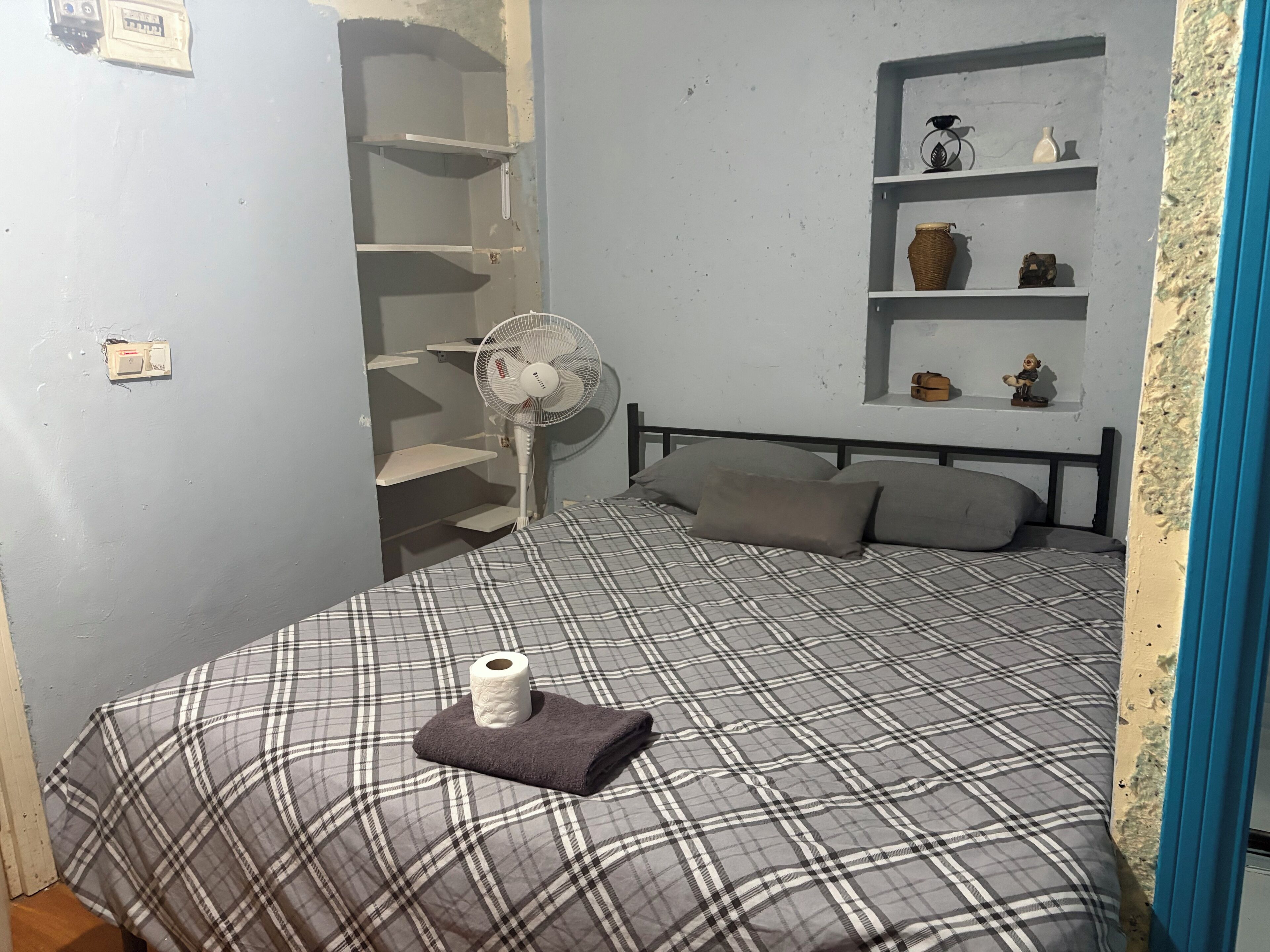 A bedroom with a bed, a fan, a shelf with decorative items, and a wardrobe.