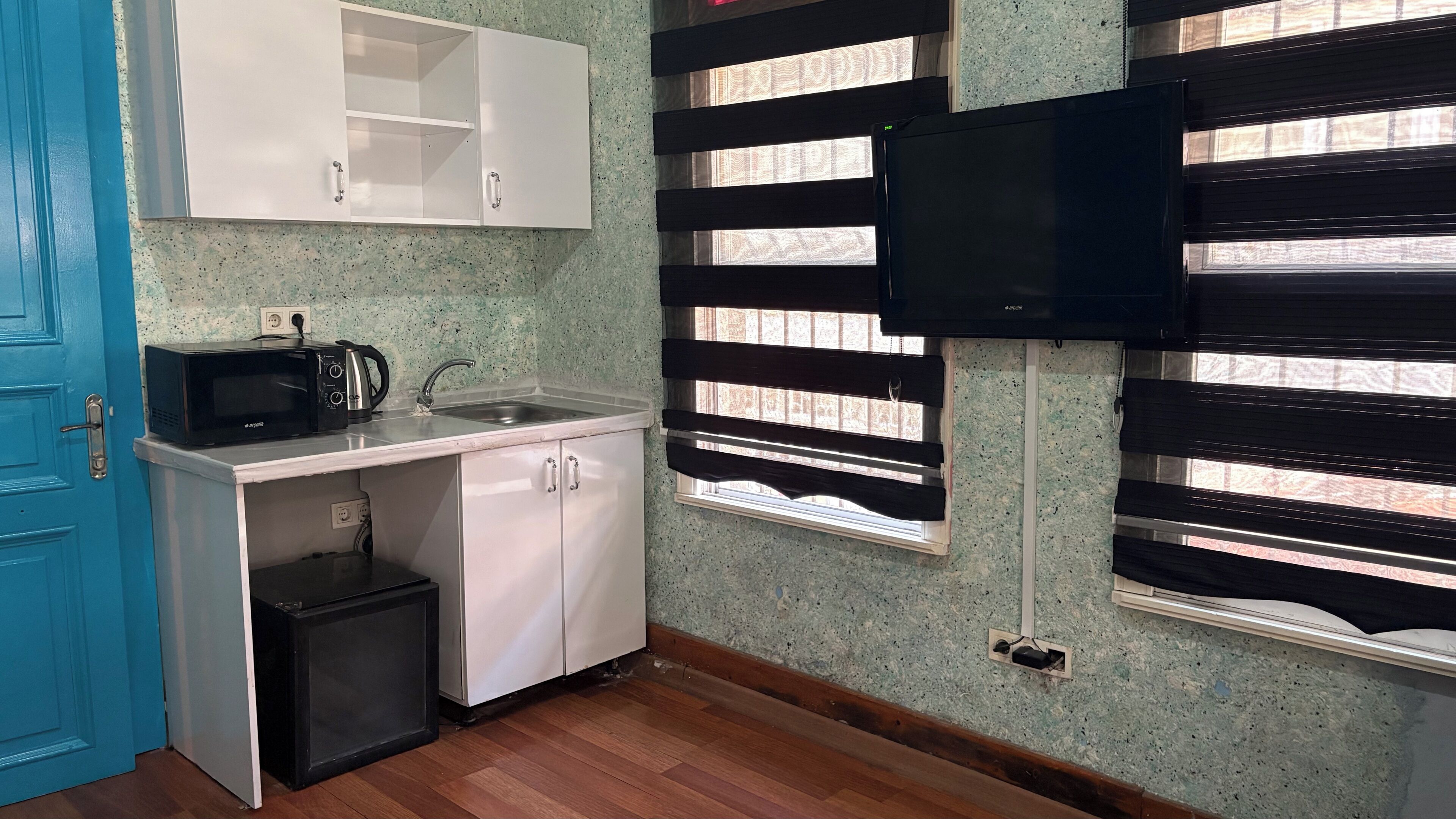 Cocina compacta con microondas, lavamanos y armarios, un refrigerador pequeño y una ventana con persianas.