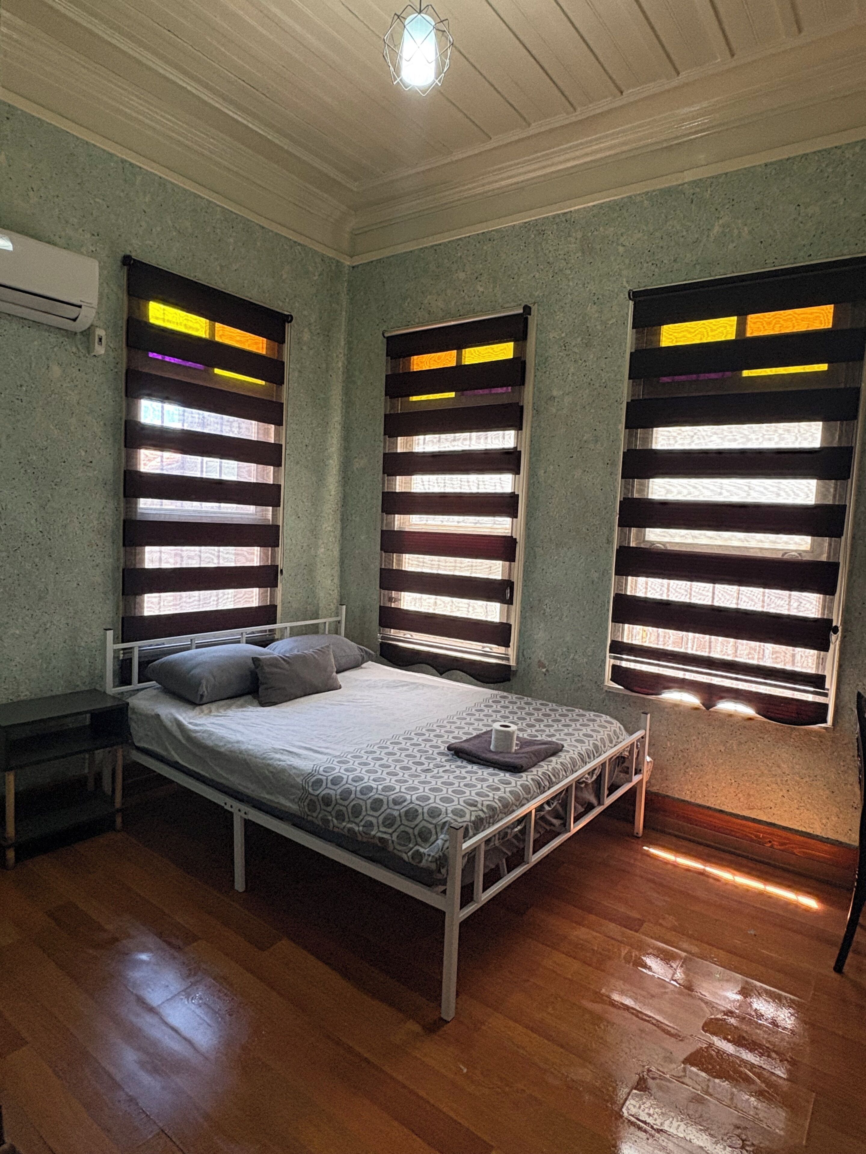 A bedroom with a single bed, a nightstand, a wooden floor, and a window with blinds.