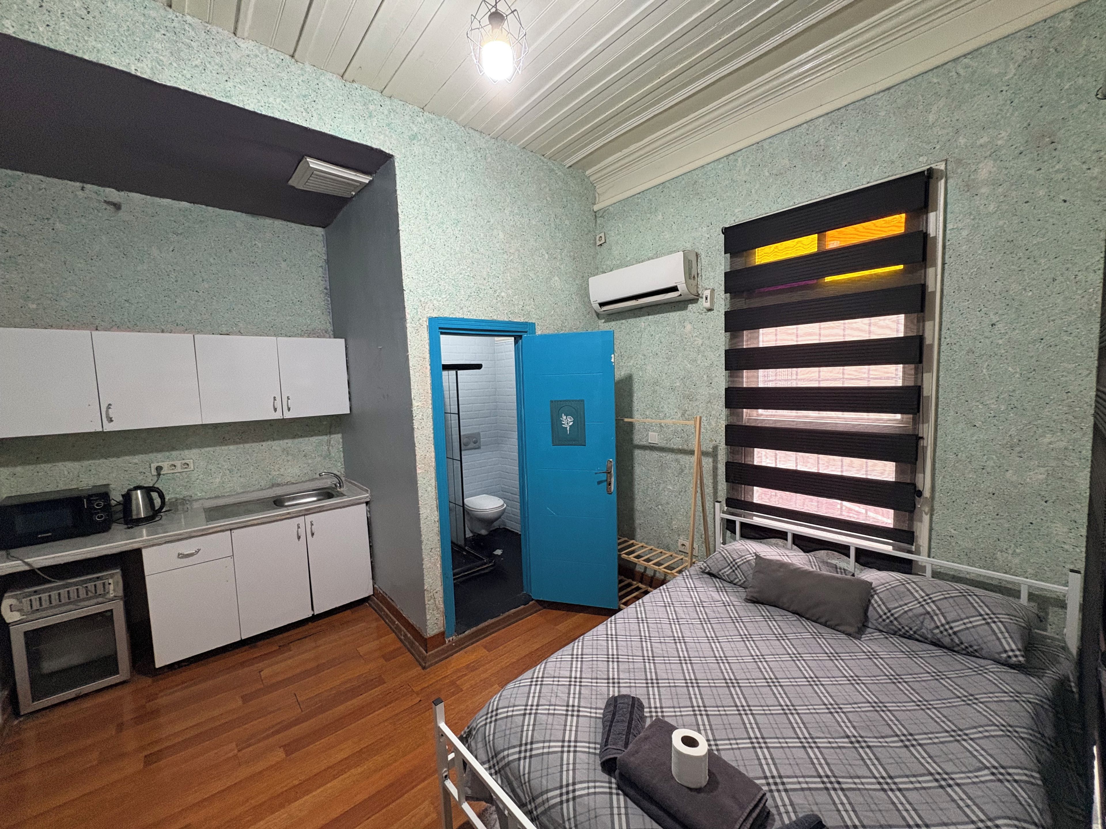 Une pièce compacte comprenant un coin cuisine, un lit avec une couette à carreaux, une porte bleue donnant sur une salle de bain et une fenêtre avec des stores.