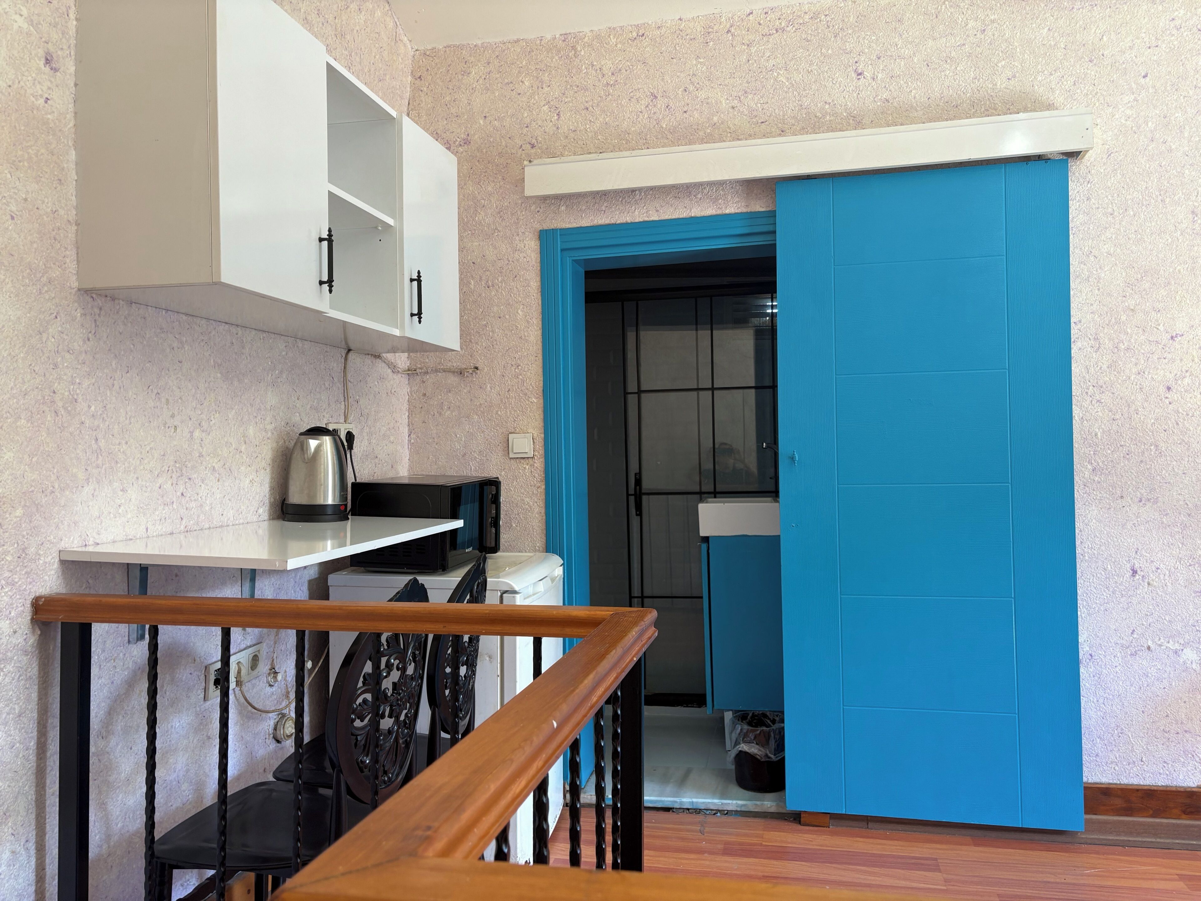A kitchen with white cabinets, a microwave, a kettle, and a blue door leading to a bathroom.