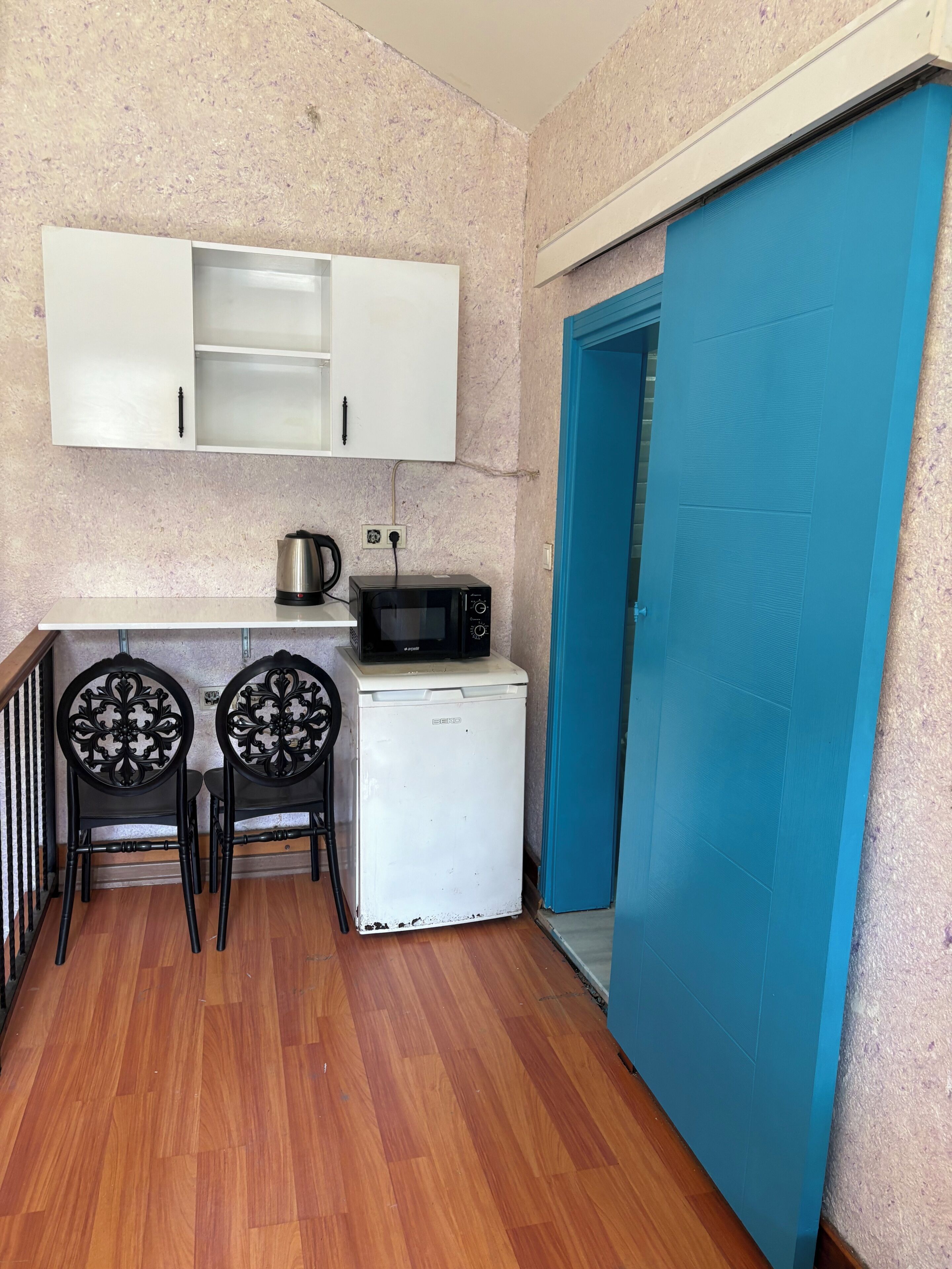 Una kitchenette compacta con microondas, hervidor de agua y un refrigerador pequeño. Hay dos sillas con respaldo decorado. Se ve una puerta azul a la derecha.